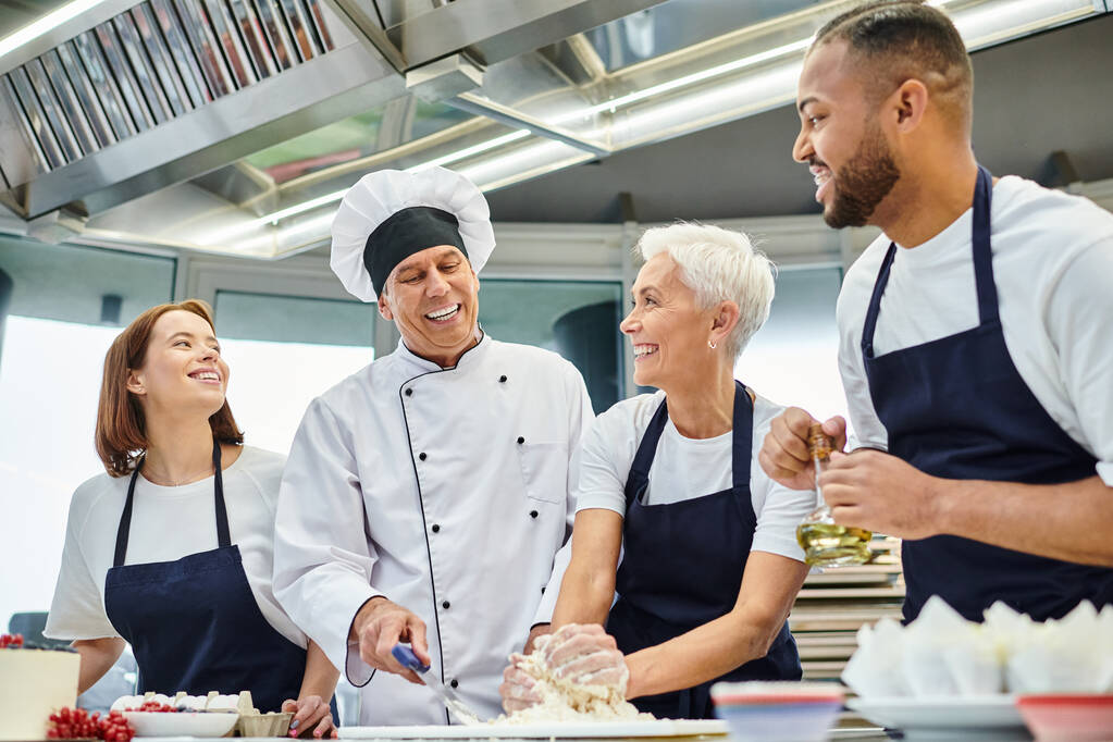 alegre cocinero jefe maduro en sombrero blanco ayudando a sus chefs multiculturales con masa, confitería - Foto, imagen