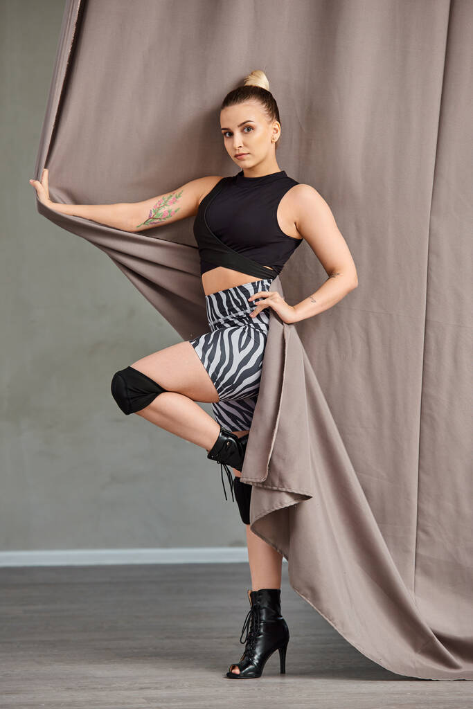 young woman in stylish clothing and high heels gracefully poses against a wall with curtain - Photo, Image