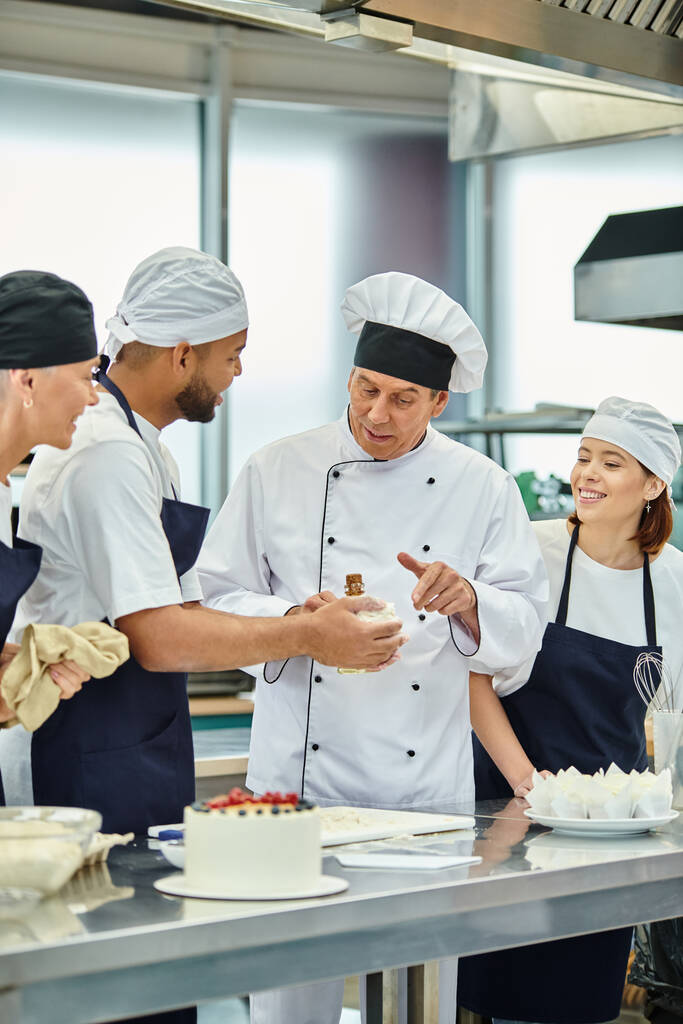 maduro guapo jefe de cocina en sombrero blanco ayudando a sus chefs interracial con masa, confitería - Foto, Imagen