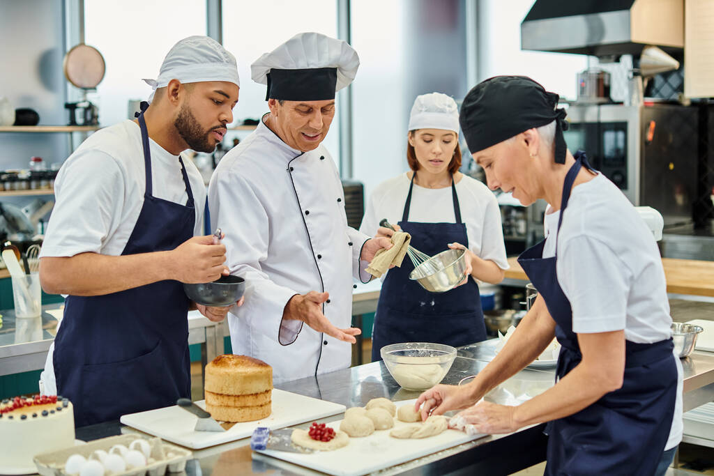 alegre cocinero jefe maduro con sus chefs multiculturales viendo mujer madura trabajando con la masa - Foto, Imagen