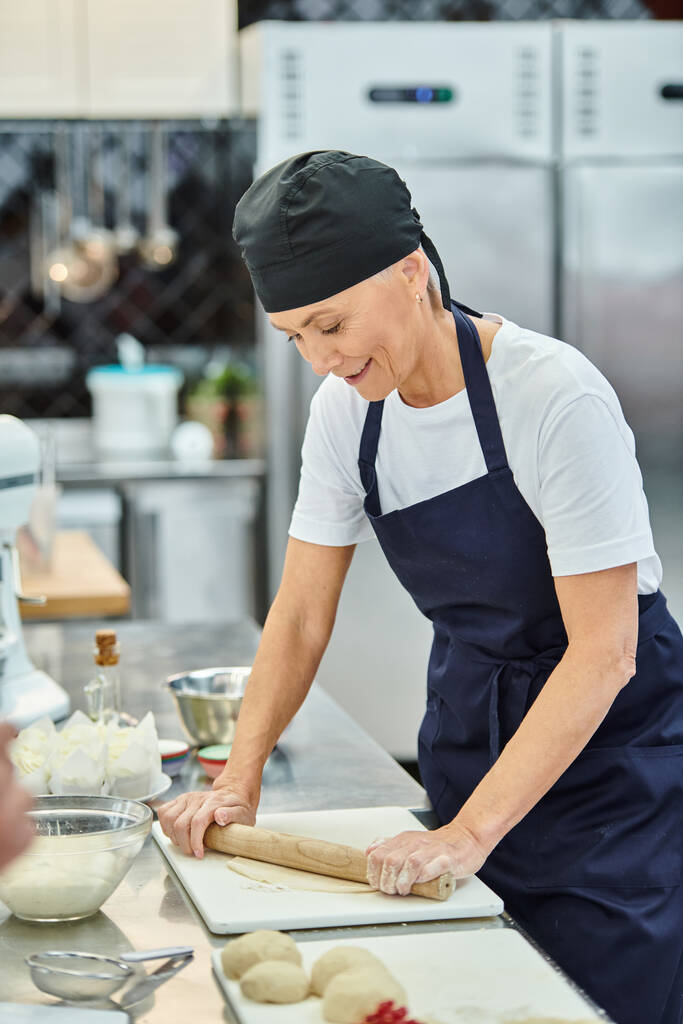 mulher bonita madura alegre no toque e avental trabalhando com massa na cozinha, confeitaria - Foto, Imagem