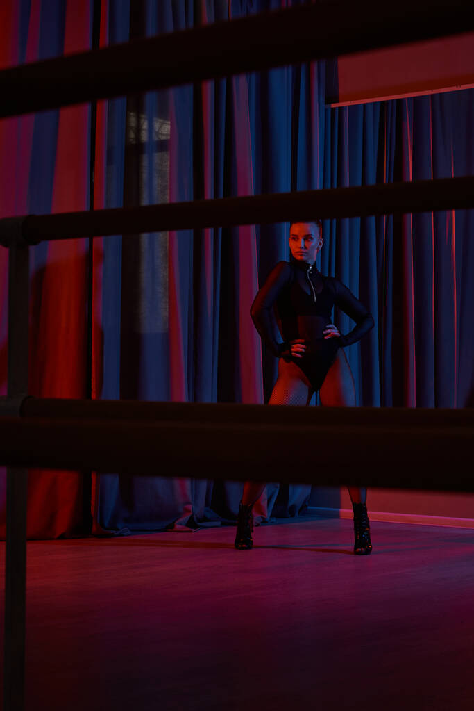 joven bailarina golpea una pose feroz en su maillot negro y medias de red, enmarcadas por la cortina - Foto, Imagen