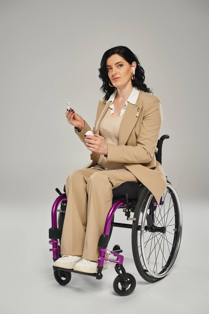 Selbstbewusste Behinderte im pastellfarbenen Anzug im Rollstuhl setzt Kopfhörer auf und blickt in die Kamera - Foto, Bild