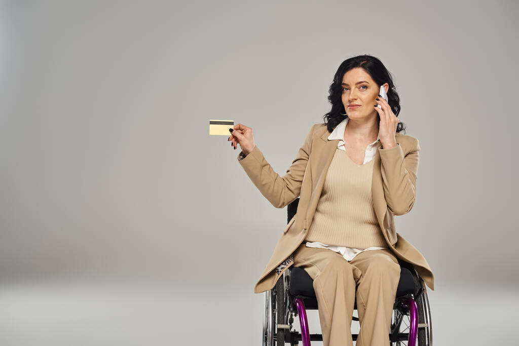 уверенная женщина-инвалид в пастельном костюме в инвалидном кресле держит кредитную карту и разговаривает по телефону - Фото, изображение