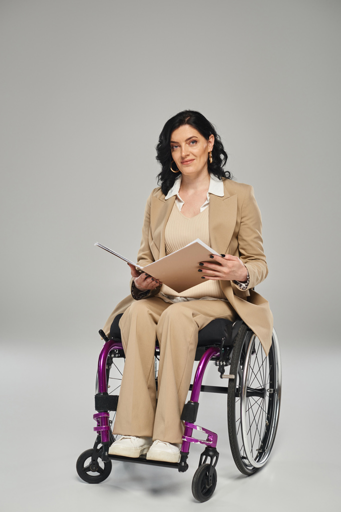 mooie brunette vrouw met een handicap zitten in een rolstoel met papierwerk en kijken naar camera - Foto, afbeelding