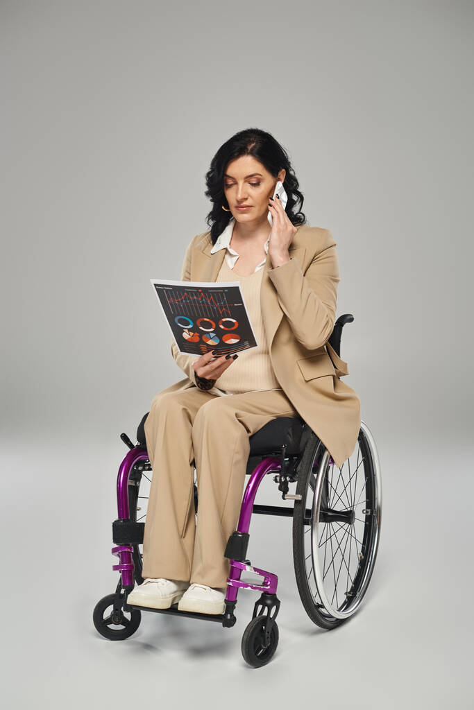 kaunis vammainen nainen tyylikkäässä puvussa keskustelee grafiikasta puhelimitse istuessaan pyörätuolissa - Valokuva, kuva