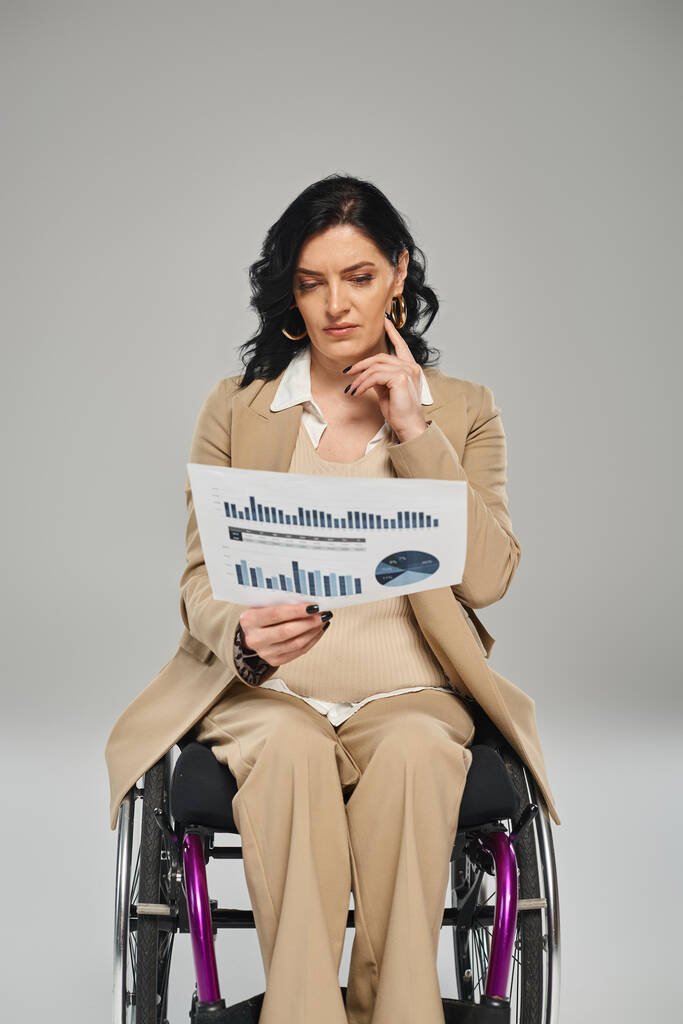 зосереджена інвалідна жінка з хвилястим волоссям у пастельному костюмі сидить у інвалідному візку і дивиться на графіку - Фото, зображення