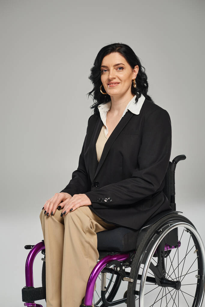 Радостная брюнетка с инвалидностью в черном блейзере сидит в инвалидном кресле и улыбается в камеру - Фото, изображение