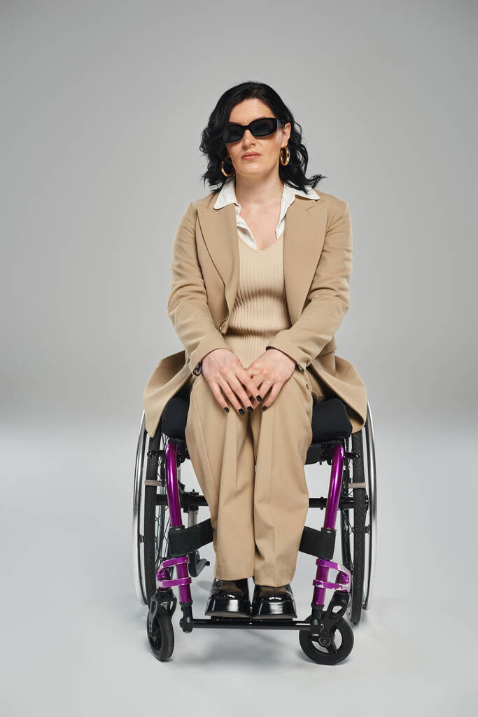atraktivní zdravotně postižená žena s vlnitými vlasy sedí na invalidním vozíku ve slunečních brýlích a dívá se do kamery - Fotografie, Obrázek