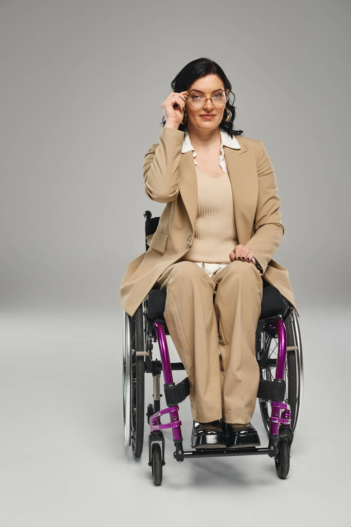 atrakcyjna brunetka niepełnosprawna kobieta w eleganckim stroju na wózku inwalidzkim z okularami patrzącymi w kamerę - Zdjęcie, obraz