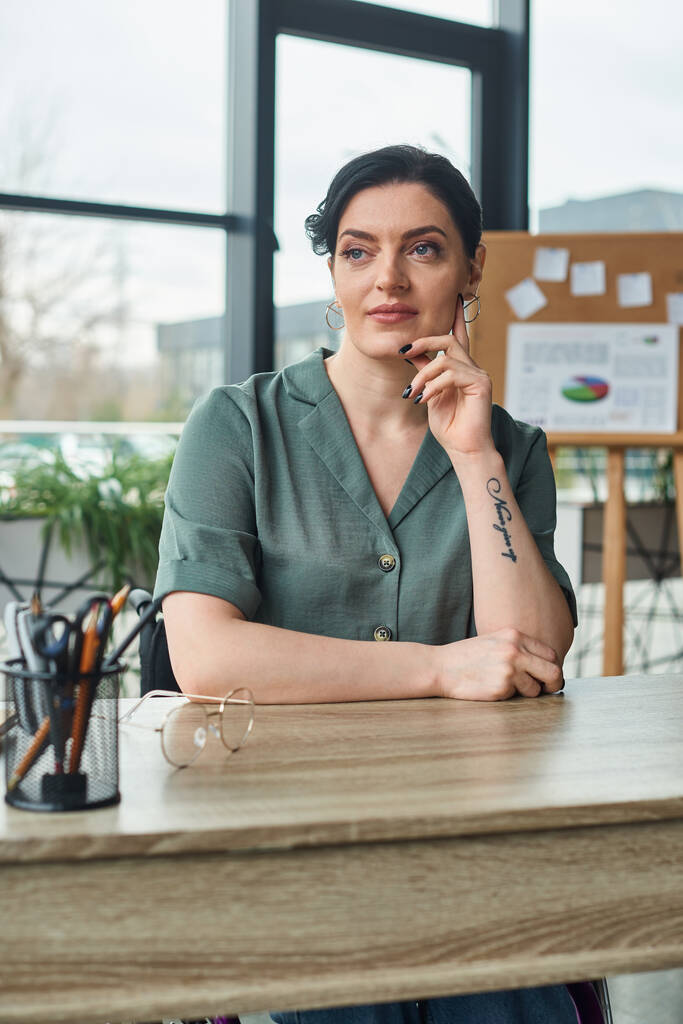 ελκυστική επιχειρηματίας με αυτοπεποίθηση σε casual ενδυμασία κάθεται στο τραπέζι και κοιτάζοντας μακριά στο γραφείο - Φωτογραφία, εικόνα