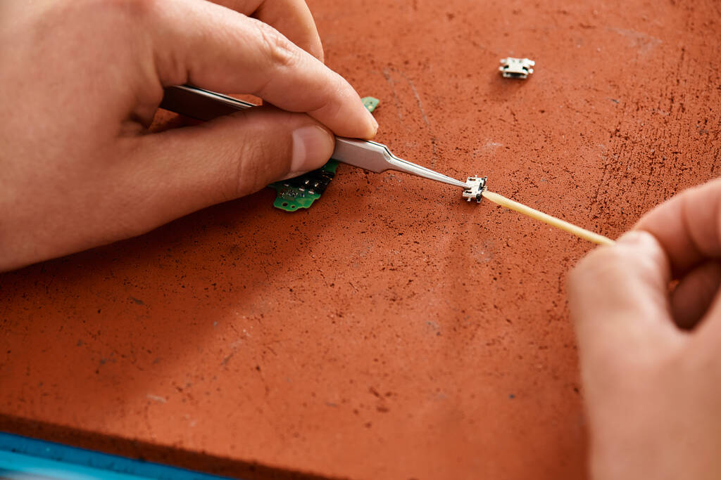 Ausgeschnittene Ansicht eines erfahrenen Reparateurs mit Pinzette beim Testen des elektronischen Chipsatzes in einer privaten Werkstatt - Foto, Bild