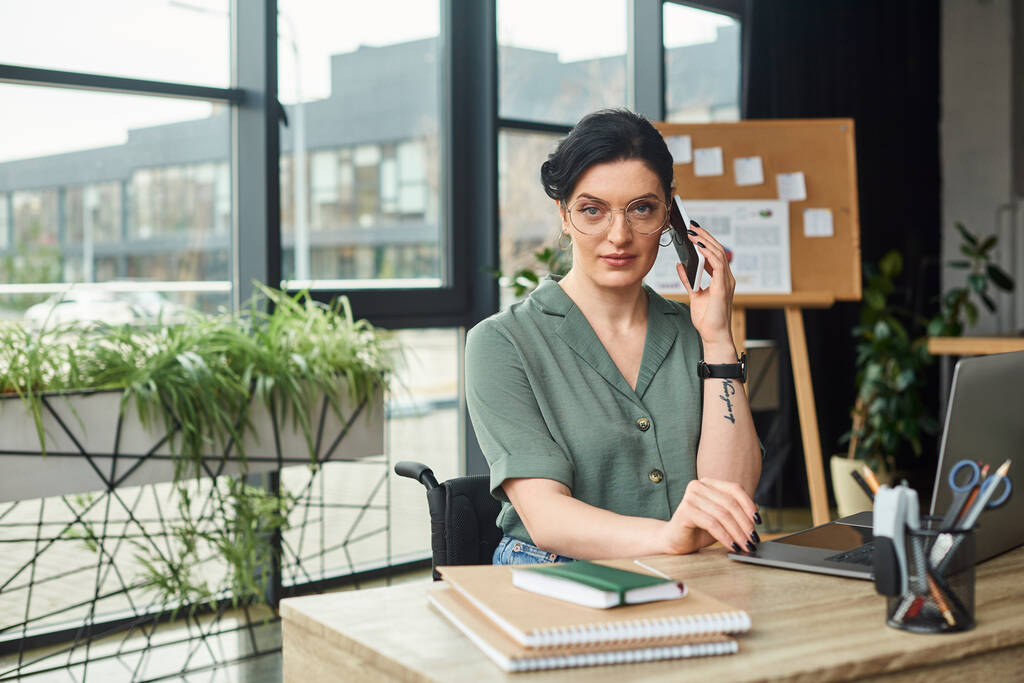 mujer discapacitada de buen aspecto pensativa en traje casual en gafas hablando por teléfono mientras está en el trabajo - Foto, Imagen