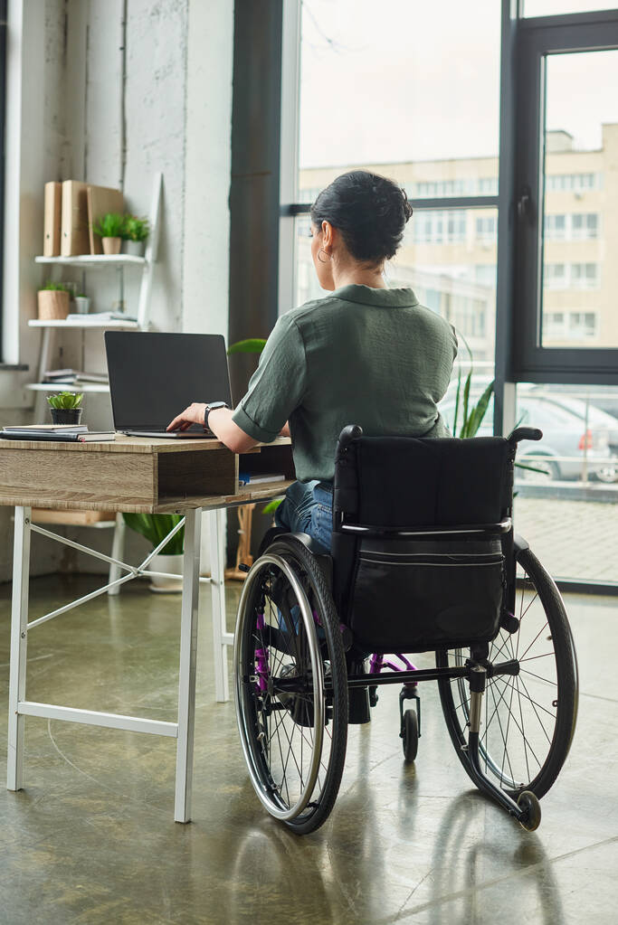посвященная брюнетка-инвалид женщина в повседневной одежде сидит в инвалидной коляске и упорно работает в офисе - Фото, изображение