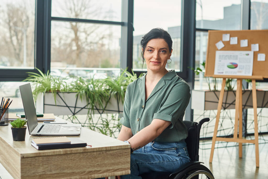 mujer atractiva alegre con discapacidad en su silla de ruedas mirando a la cámara durante el trabajo en la oficina - Foto, Imagen