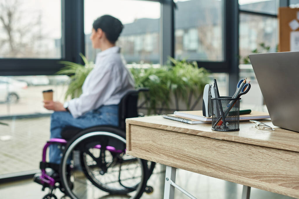 фокус на столе с канцелярскими принадлежностями с размытой женщиной-инвалидом в инвалидной коляске на заднем плане в офисе - Фото, изображение