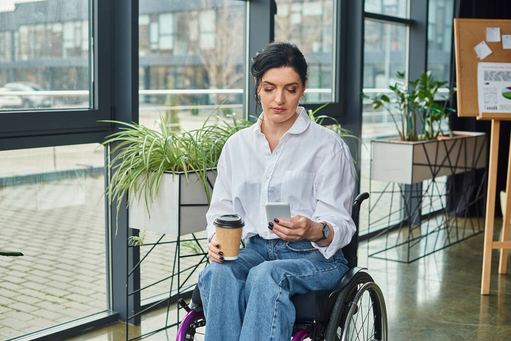 atractiva morena mujer discapacitada en elegante atuendo en silla de ruedas sosteniendo café y teléfono inteligente - Foto, imagen