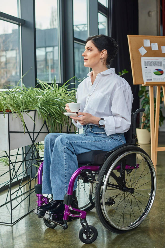 atraktivní oddaná podnikatelka se zdravotním postižením v invalidním vozíku drží šálek kávy a dívá se jinam - Fotografie, Obrázek