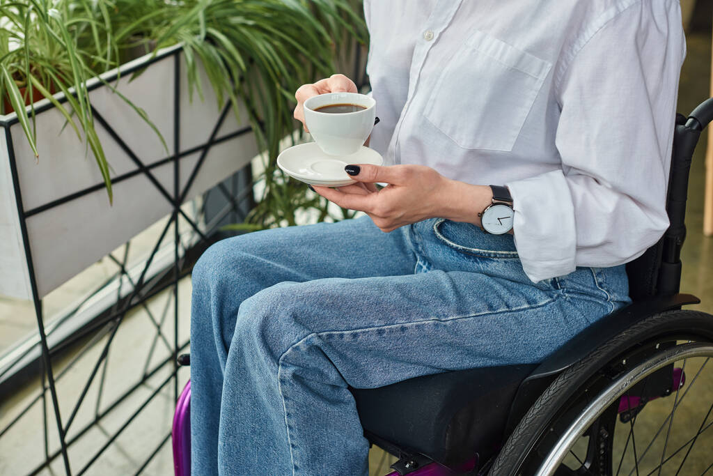 καλλιεργούμενη άποψη της γυναίκας επιχειρηματία με αναπηρία κινητικότητας σε αναπηρικό αμαξίδιο κρατώντας φλιτζάνι καφέ στο γραφείο - Φωτογραφία, εικόνα