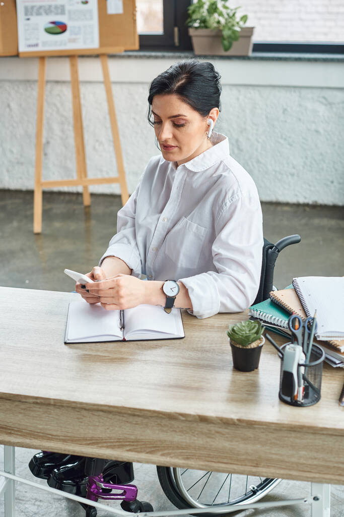 attraente donna disabile in elegante abbigliamento con le cuffie guardando il suo telefono mentre in ufficio - Foto, immagini