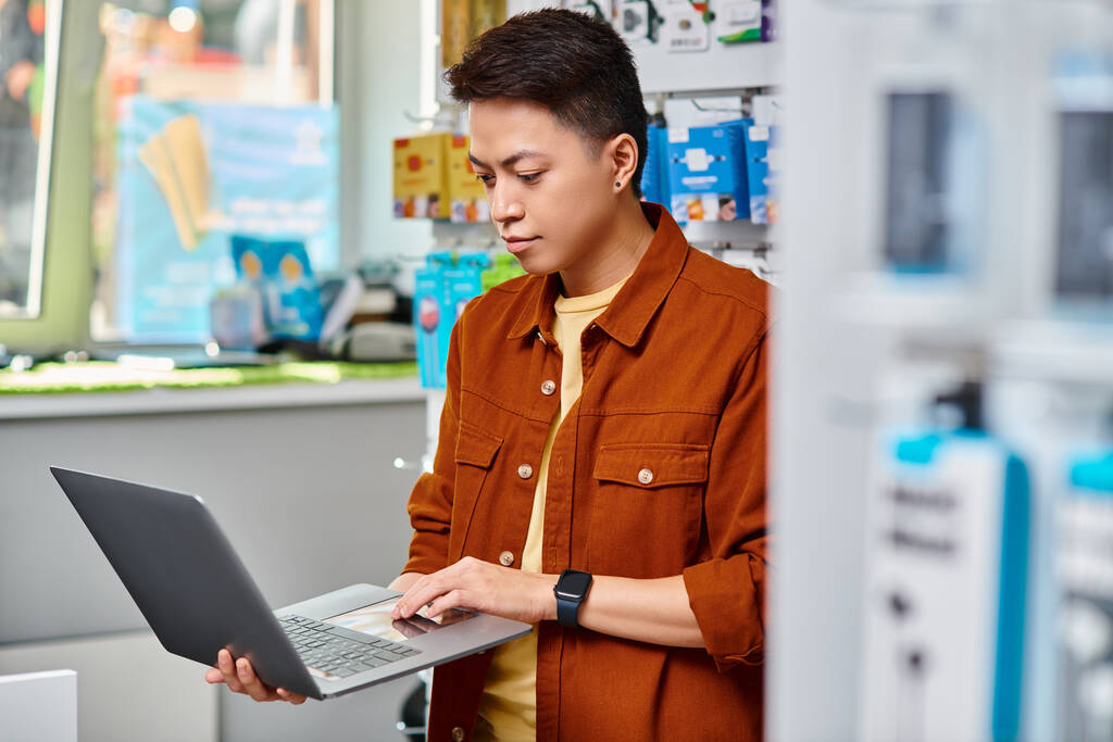 концентрований азіатський чоловік використовує ноутбук, стоячи в приватному магазині електроніки, малий бізнес - Фото, зображення