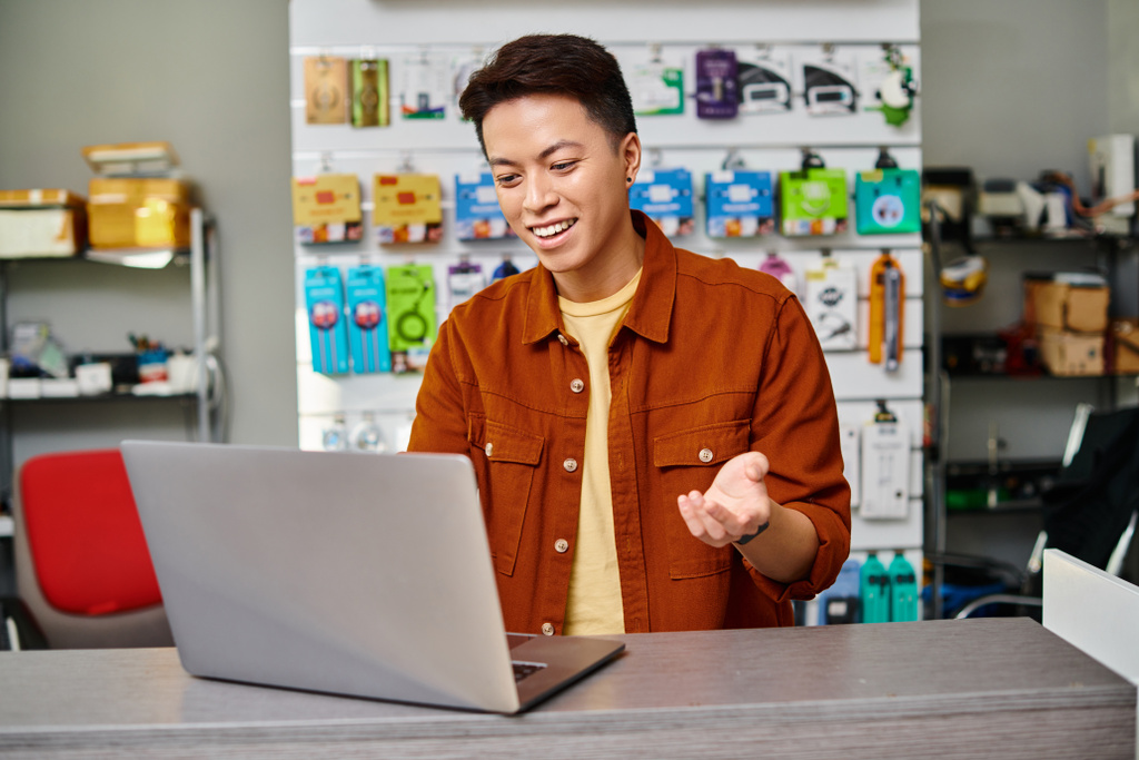 θετική ασιατική πωλητής gesturing κατά τη διάρκεια της online πώλησης σε φορητό υπολογιστή στον πάγκο στο δικό του κατάστημα ηλεκτρονικών ειδών - Φωτογραφία, εικόνα