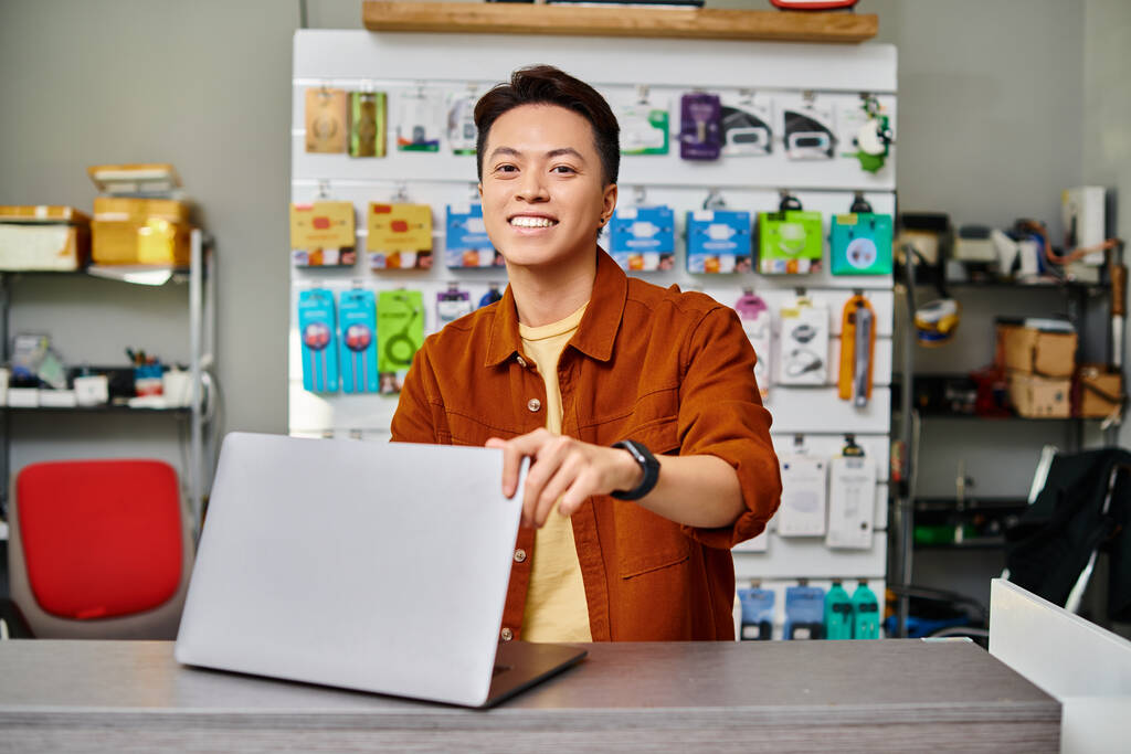 alegre asiático electrónica tienda propietario mirando cámara cerca de portátil en contador en privado tienda - Foto, Imagen