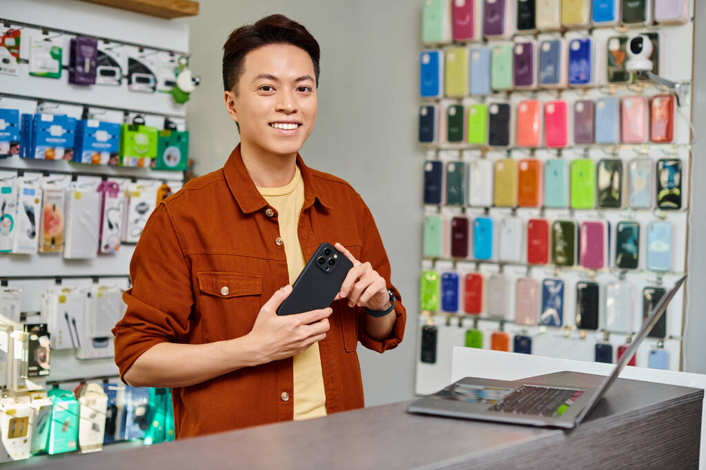 電子工学の店のカウンターのラップトップの近くのカメラを見ているスマートフォンが付いている陽気なアジアの男 - 写真・画像