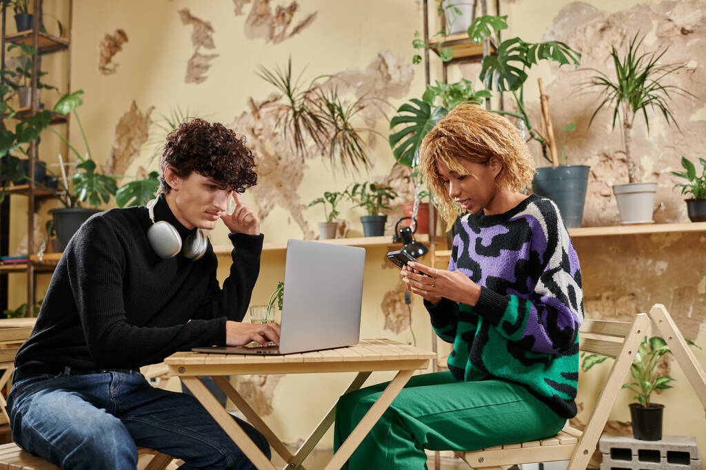 couple jeune et diversifié en utilisant des appareils tout en étant assis àtable dans un café végétalien rempli de plantes - Photo, image
