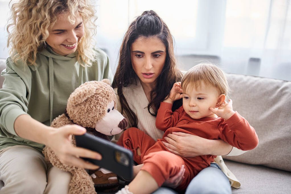 caring lgbt couple on sofa with their baby girl looking at phone holding teddy bear, family concept - Photo, Image