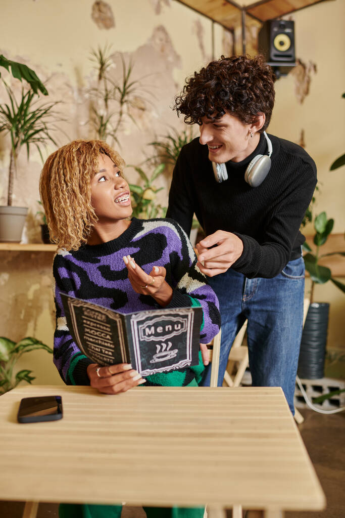позитивный и молодой мужчина и женщина в повседневной одежде читают меню в веганском кафе, межрасовая пара - Фото, изображение