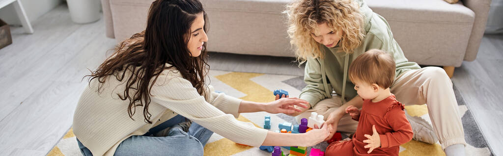 χαρούμενο ζευγάρι λεσβιών που παίζει με το κοριτσάκι τους με παιχνίδια στο πάτωμα στο σπίτι, οικογενειακή ιδέα, πανό - Φωτογραφία, εικόνα
