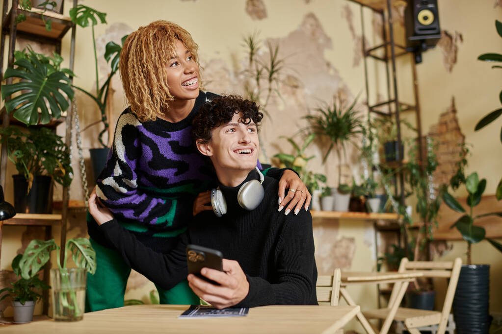 mujer afro-americana positiva con el pelo rizado abrazando hombre joven con teléfono inteligente en la cafetería vegana - Foto, imagen