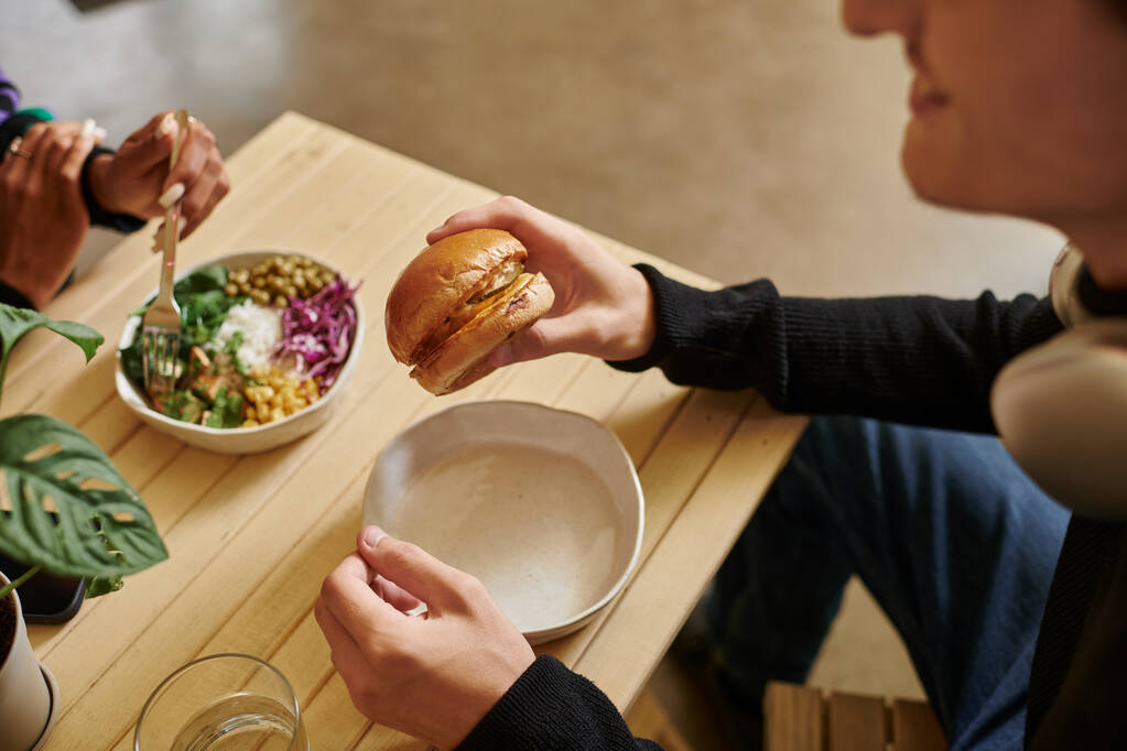 καλλιεργούνται νεαρός άνδρας απολαμβάνοντας χορτοφαγικό γεύμα, ενώ κρατώντας burger με τόφου κοντά θολή γυναίκα τρώει σαλάτα - Φωτογραφία, εικόνα