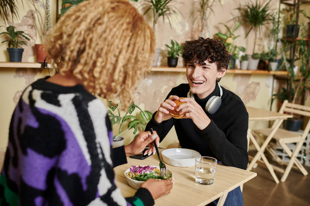 счастливый кудрявый человек ест бургер с тофу рядом с африканской американской подружкой наслаждаясь салатом в веганском кафе - Фото, изображение
