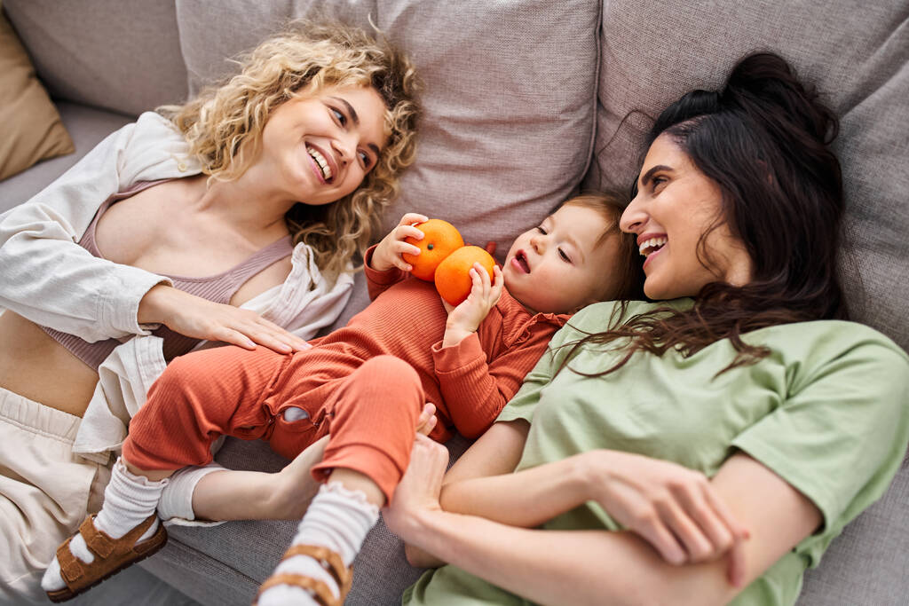 feliz pareja lgbt acostado en el sofá con su bonita niña sosteniendo mandarinas, concepto de familia - Foto, imagen