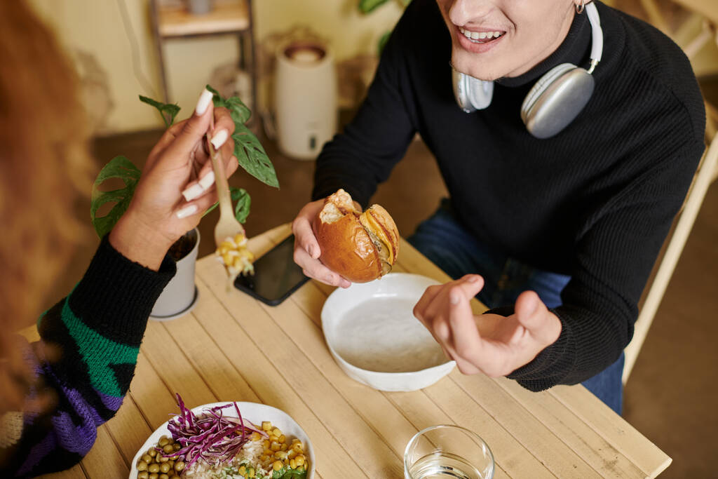 recortado disparo de hombre feliz en auriculares inalámbricos comer hamburguesa de tofu vegano cerca de la mujer en la cafetería - Foto, Imagen