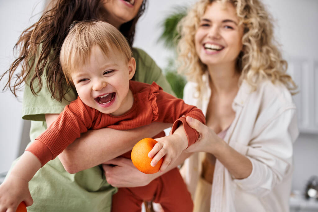 cheerful lesbian couple having fun together with their cute baby girl holding tangerines, family - Photo, Image