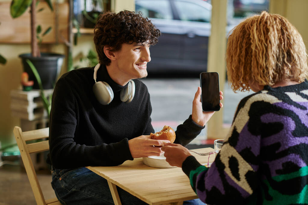 szczęśliwy kręcone człowiek z bezprzewodowych słuchawek gospodarstwa tofu burger i smartfon w wegańskiej kawiarni - Zdjęcie, obraz