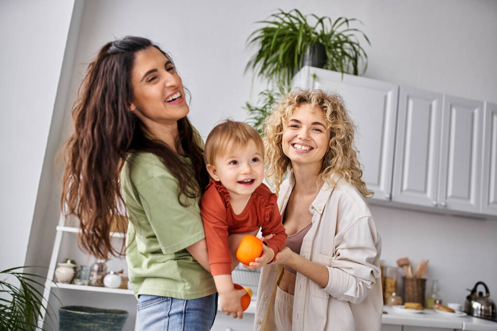 alegre pareja de lesbianas divirtiéndose junto con su lindo bebé niña sosteniendo mandarinas, familia - Foto, Imagen