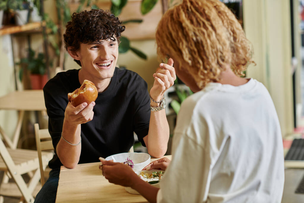 hombre sonriente y rizado comiendo hamburguesa de tofu cerca de chica afroamericana disfrutando de ensalada en la cafetería vegana - Foto, imagen