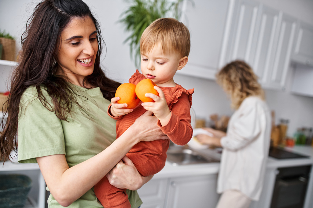 focus on brunette woman with daughter holding tangerines with her blurred blonde partner on backdrop - Photo, Image