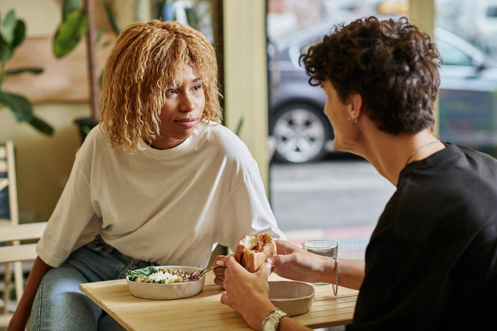 молода афроамериканка з кучерявим волоссям їсть салатну миску біля розмитого хлопця у веганському кафе - Фото, зображення