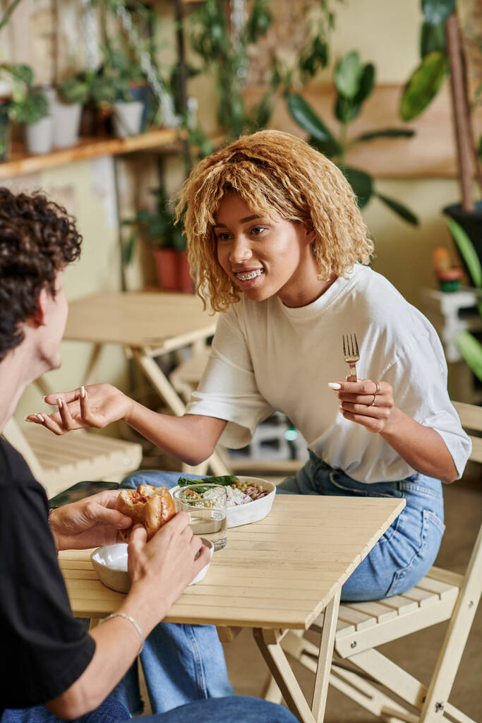 счастливая африканская американская девушка болтает с кудрявым парнем возле вегетарианской еды в веганском кафе - Фото, изображение