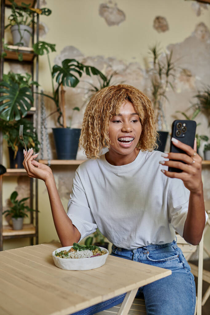 збуджена темношкіра молода жінка з брекетами, що тримає виделку біля веганського салату і дивиться на смартфон - Фото, зображення