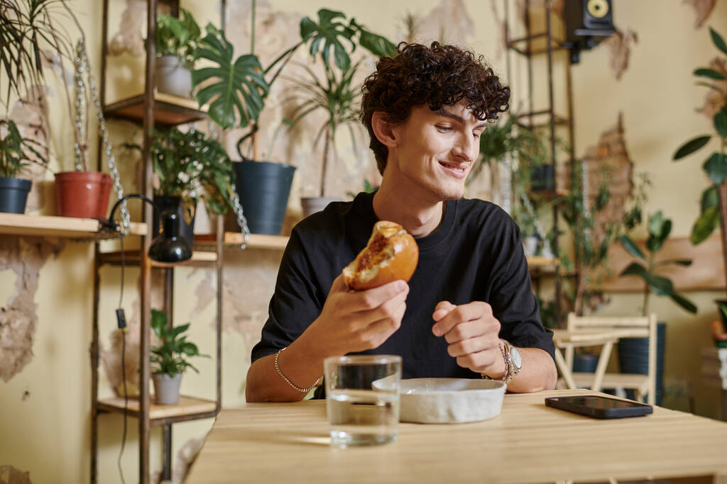 счастливый и молодой кудрявый мужчина держит бургер с тофу и смотрит на смартфон на столе в веганском кафе - Фото, изображение