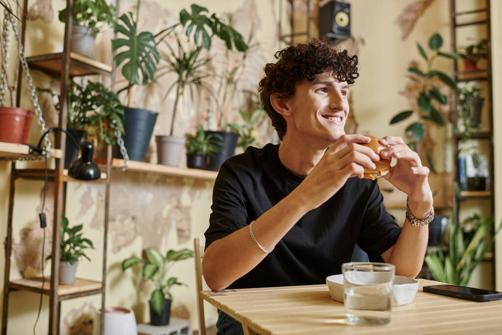 onnellinen nuori mies pitelee tofu-hampurilaista ja katsoo pois hymyillen vegaanisessa kahvilassa, nautinto - Valokuva, kuva