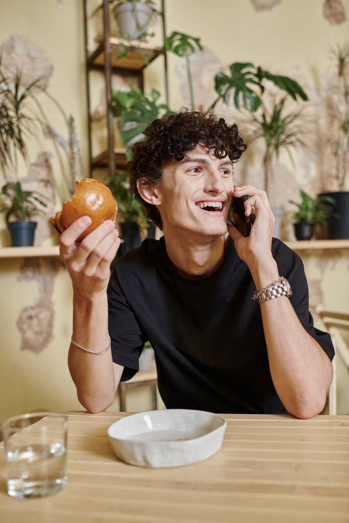 позитивний молодий кучерявий чоловік тримає рослинний бургер на тофу і розмовляє на смартфоні у веганському кафе - Фото, зображення