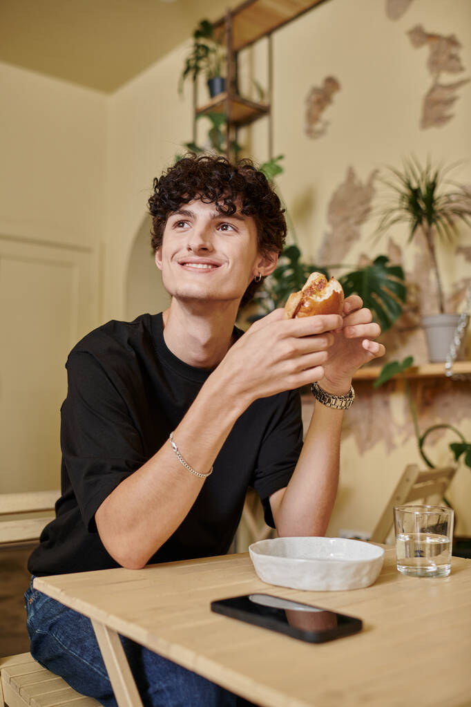joven feliz y rizado sosteniendo hamburguesa de tofu orgánica y sonriendo en la planta llena de café vegano - Foto, Imagen