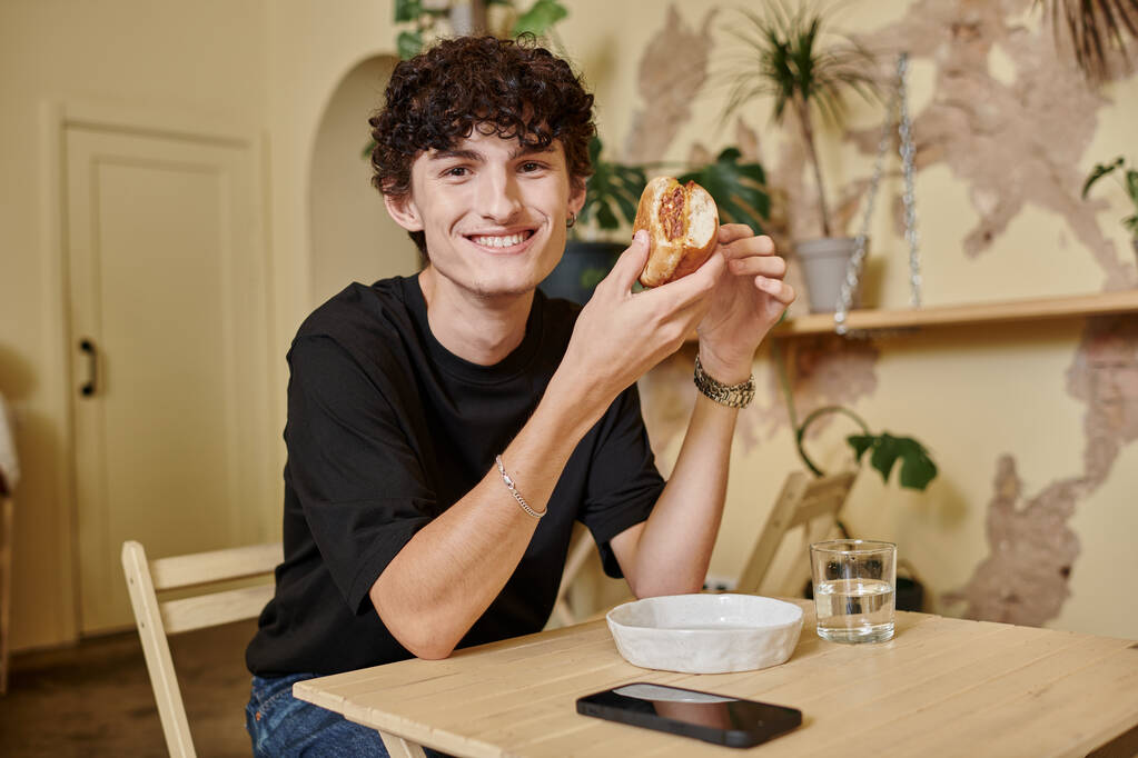χαρούμενα και σγουρά νεαρός άνδρας που κατέχουν βιολογικά tofu burger και χαμογελώντας σε φυτό γεμάτο vegan καφέ - Φωτογραφία, εικόνα